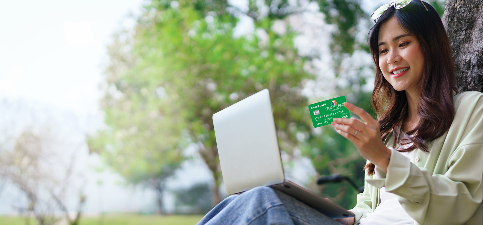 woman sitting against tree on laptop holding her debit card