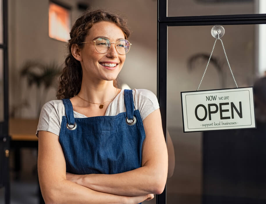 A young woman wearing an apron standing next to a "We're Open" sign.